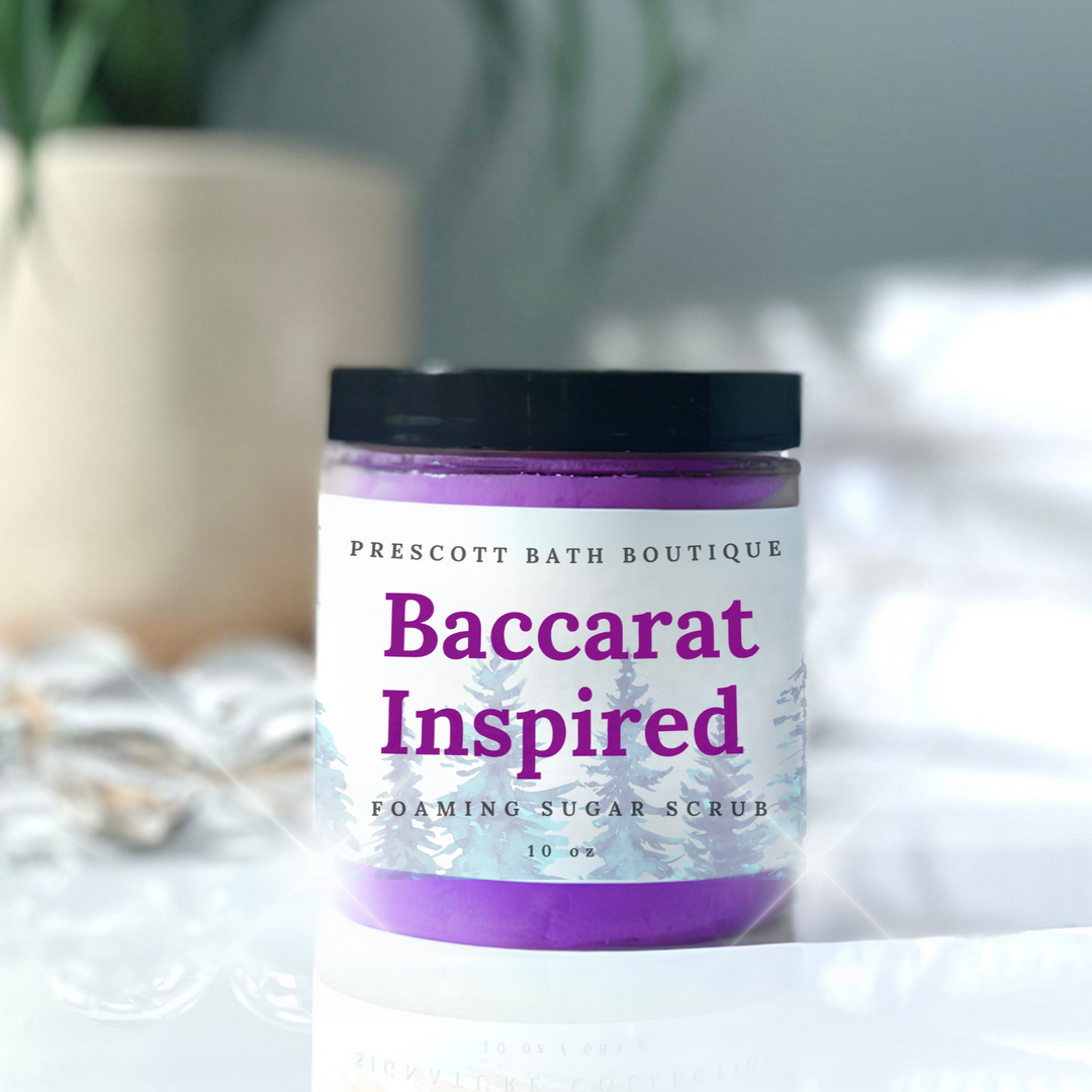 Jar of 'Baccarat Inspired' foaming sugar scrub from Prescott Bath Boutique on a white surface with a blurred plant in the background.