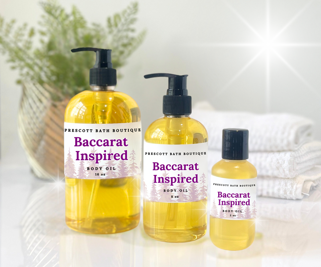 Three bottles of 'Baccarat Inspired' body oil from Prescott Bath Boutique on a white surface with a blurred background.
