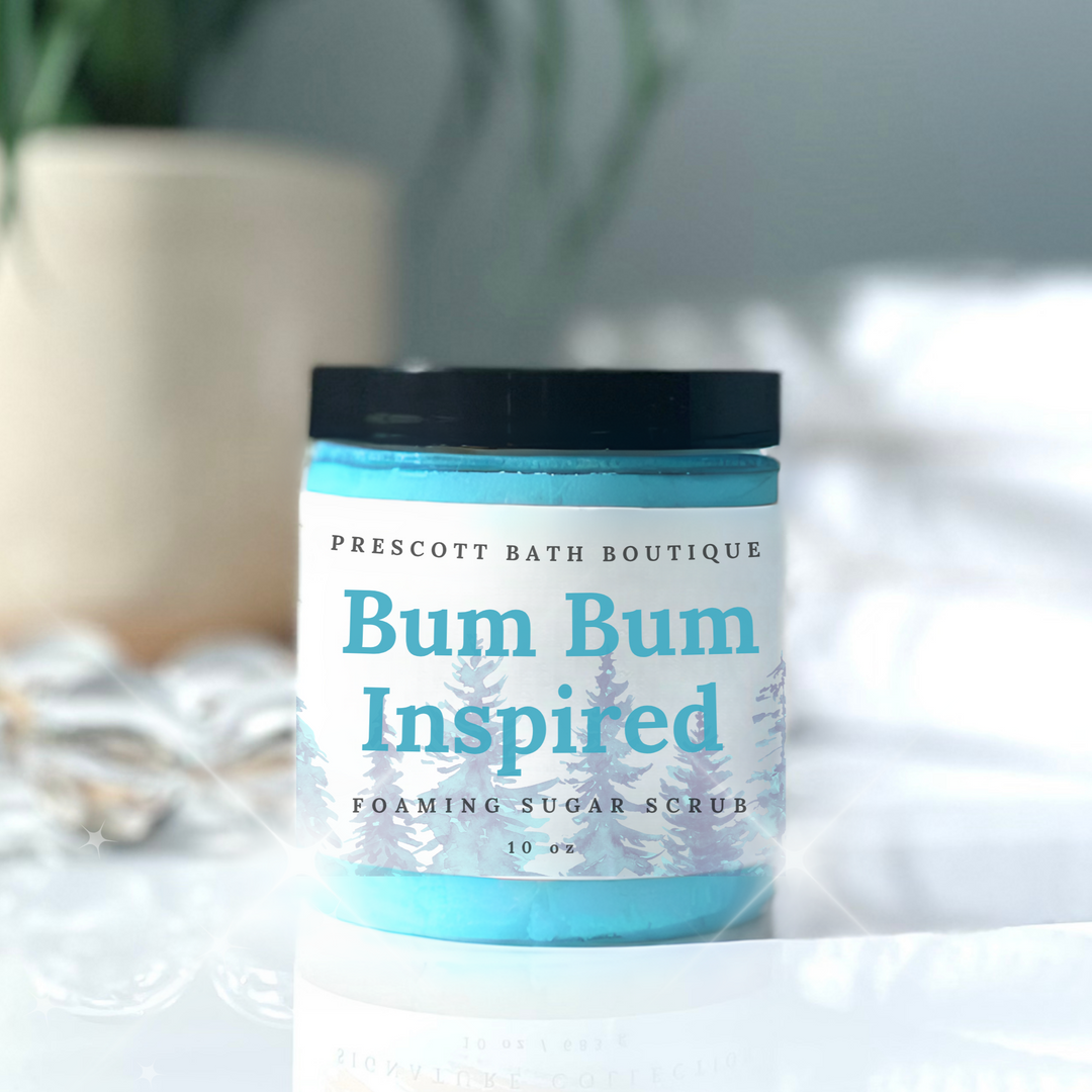 Jar of 'Bum Bum Inspired' foaming sugar scrub from Prescott Bath Boutique on a white surface with a blurred plant in the background.