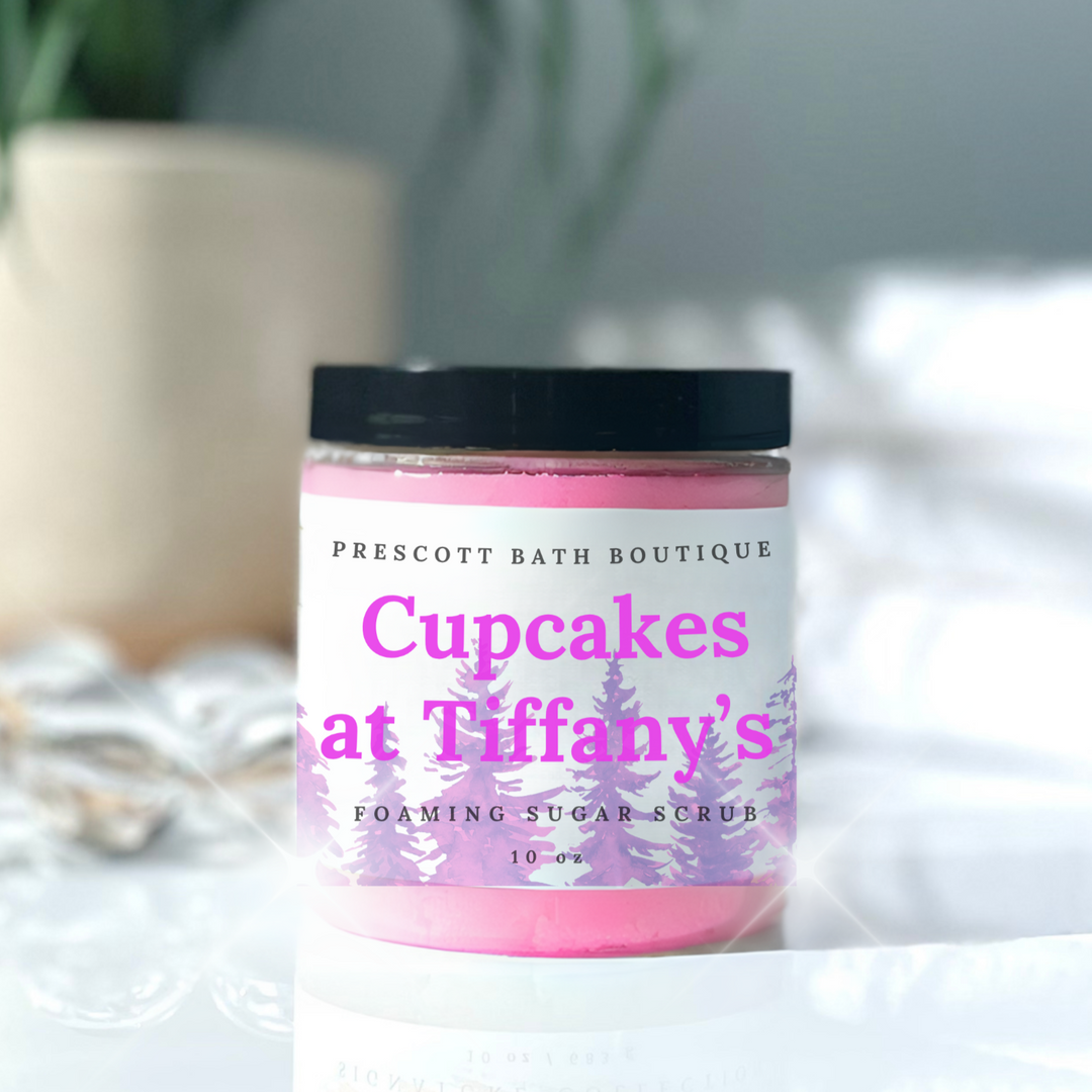 Jar of 'Cupcakes at Tiffany's' foaming sugar scrub from Prescott Bath Boutique on a white surface with a blurred plant in the background.