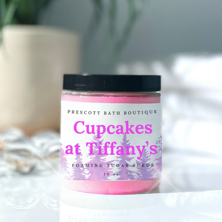 Jar of 'Cupcakes at Tiffany's' foaming sugar scrub from Prescott Bath Boutique on a white surface with a blurred plant in the background.