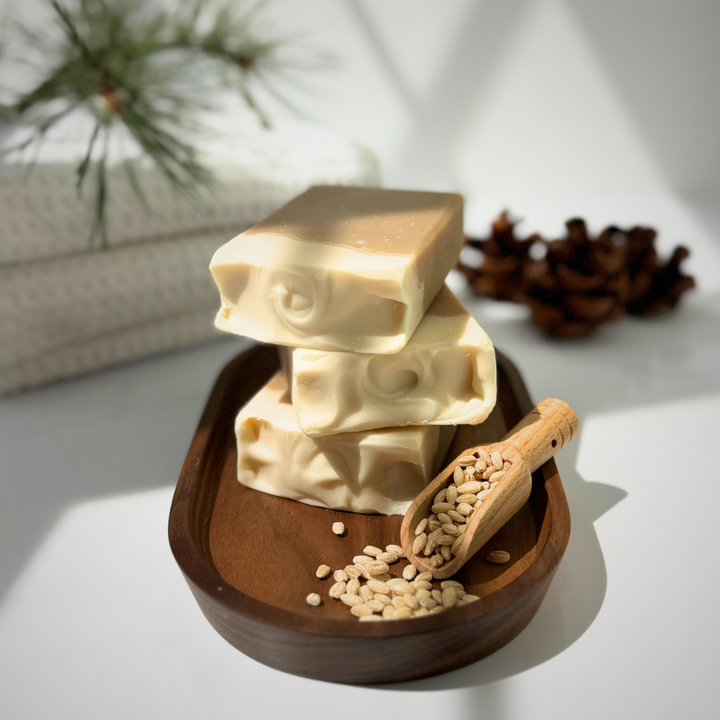 Stack of soap bars on a wooden tray with a wooden scoop containing seeds, on a light background.