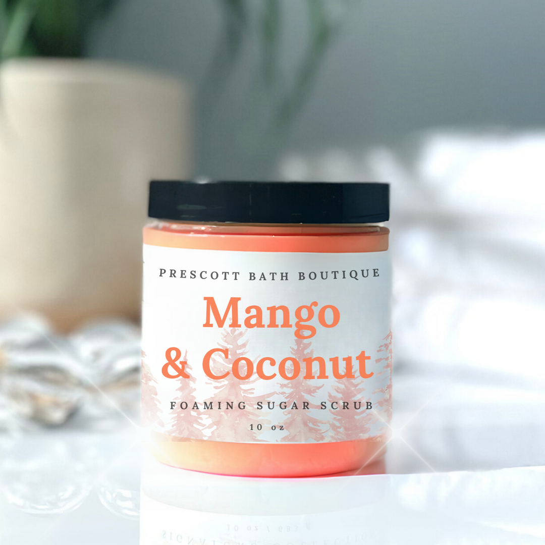 Jar of Mango & Coconut foaming sugar scrub from Prescott Bath Boutique on a white surface with a blurred plant in the background.