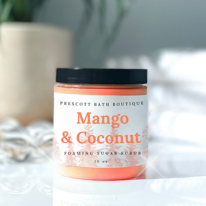 Jar of Mango & Coconut foaming sugar scrub from Prescott Bath Boutique on a white surface with a blurred plant in the background.