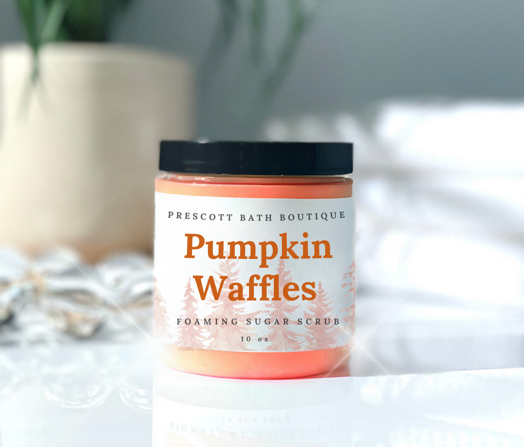 Jar of 'Pumpkin Waffles' foaming sugar scrub from Prescott Bath Boutique on a white surface with a blurred plant in the background.