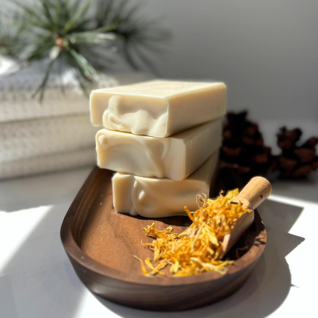Three bars of soap stacked on a wooden tray with dried flowers, against a neutral background.