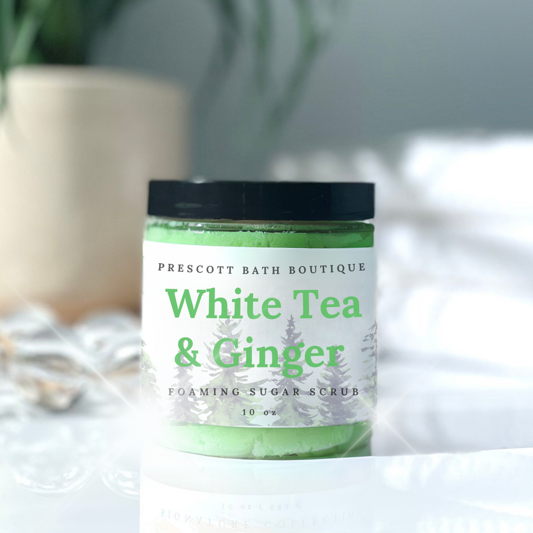 Jar of White Tea & Ginger foaming sugar scrub from Prescott Bath Boutique on a white surface with a blurred plant in the background.